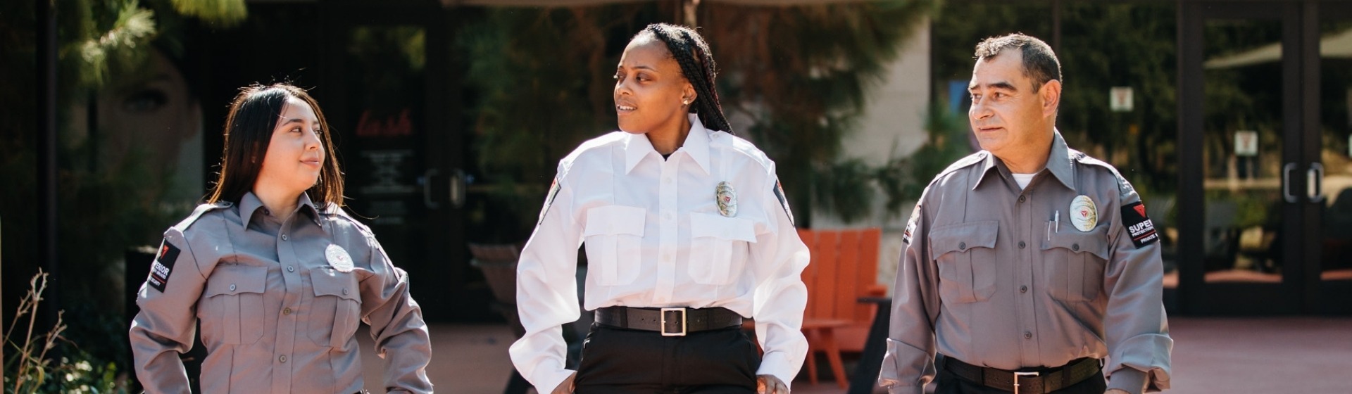 Three security guards walking in a line wearing white shirts and gray pants with a building and trees in the background. The image serves as a background for the hero section of the Office page.