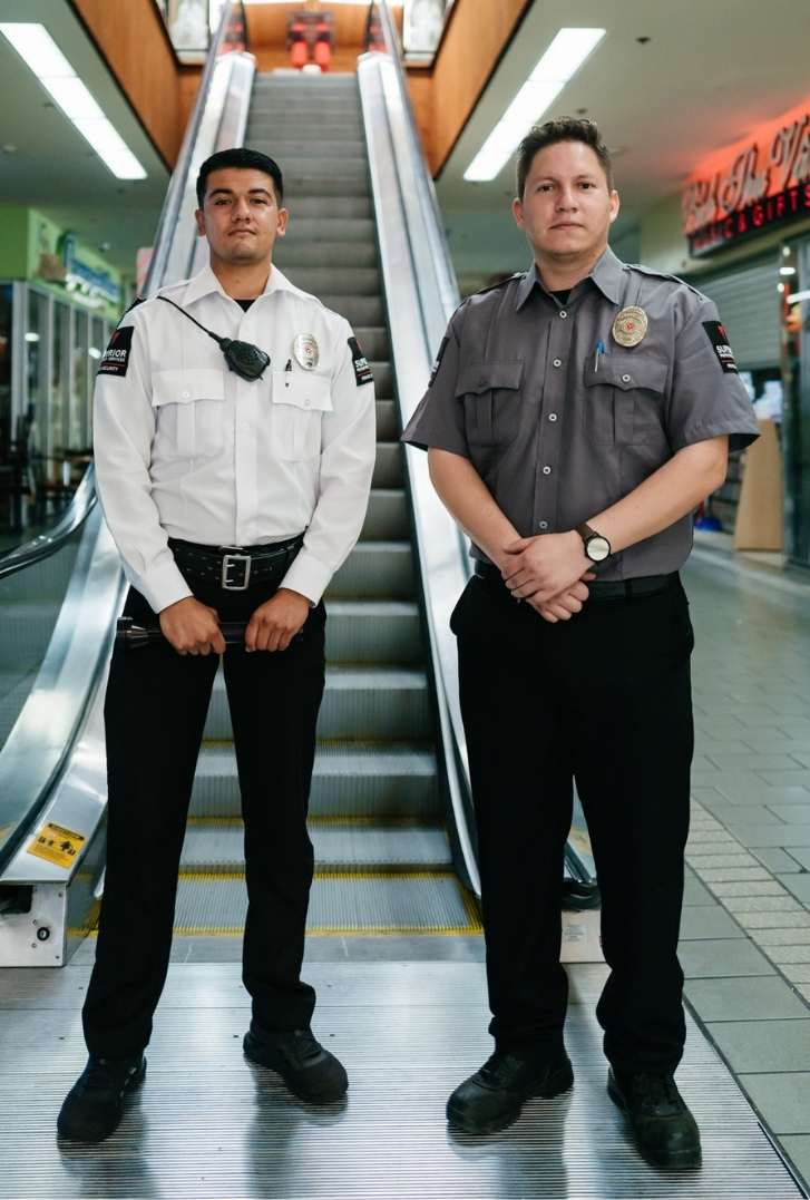 Two Superior Protection Services security personnel in different uniforms standing in front of an escalator in a mall with a store sign visible in the background.
