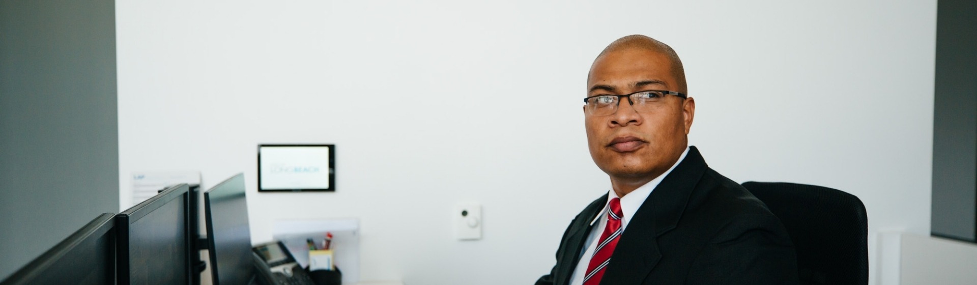 A Superior Protection Services specialist in a black suit and red tie sitting in an office in front of a desk with a computer monitor and keyboard, with a white wall in the background. The image serves as a background for the hero section of the contact us page.