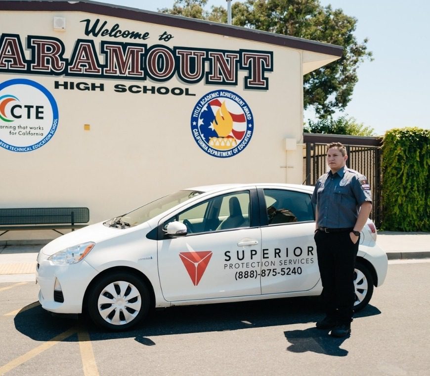 A uniformed security guard stands next to a Superior Protection Services patrol car outside Paramount High School, providing trusted security services
