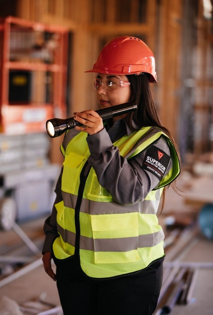 A professional female security officer in a high-visibility vest and hard hat inspects a construction site with a flashlight, ensuring top-notch security services