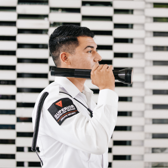 A guard in a white uniform holding a black flashlight with a white wall with black horizontal lines in the background. Security guard services in Riverside County