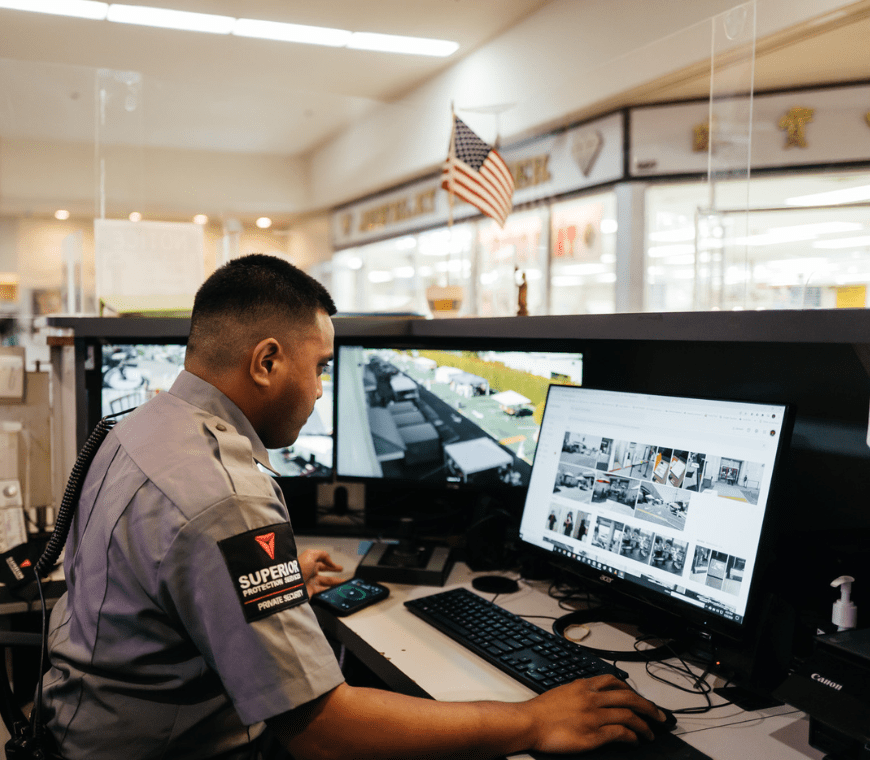A Superior Protection Services CCTV analyst in a gray uniform, sitting at a desk with multiple computer monitors displaying security camera footage.